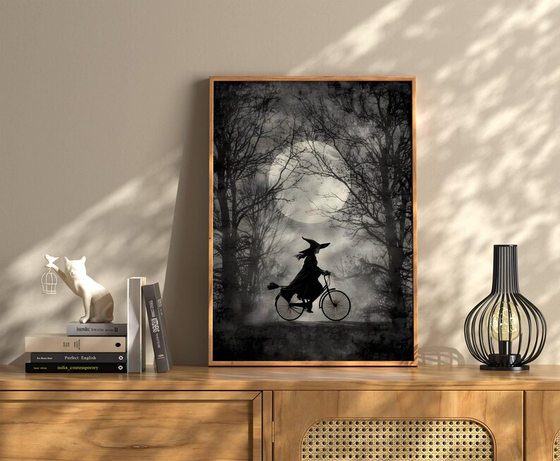 K&ouml;nnte beinhalten: Gerahmter Schwarz-Wei&szlig;-Kunstdruck mit einer Hexe, die vor einem Vollmond Fahrrad f&auml;hrt. Das Bild hat ein gruseliges Halloween-Thema mit kahlen B&auml;umen und einem nebligen Hintergrund. Der Druck wird auf einem Holzschrank ausgestellt.