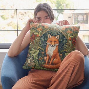 May include: A woman is sitting on a blue armchair and holding a throw pillow with a fox illustration. The pillow has a green, brown, and orange floral pattern. The fox is orange and white and is sitting in a green forest.