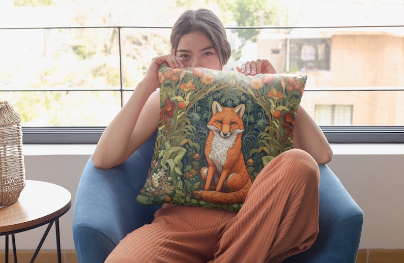 May include: A woman is sitting on a blue armchair and holding a throw pillow with a fox illustration. The pillow has a green, brown, and orange floral pattern. The fox is orange and white and is sitting in a green forest.