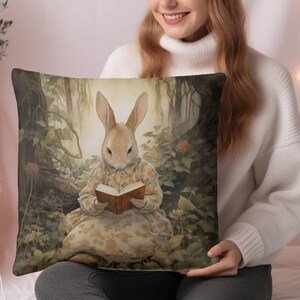 K&ouml;nnte beinhalten: Ein Dekokissen mit einer skurrilen Illustration eines Kaninchens, das ein Kleid tr&auml;gt und in einer Waldlandschaft ein Buch liest. Das Kissen hat eine braune, gr&uuml;ne und gelbe Farbgebung.