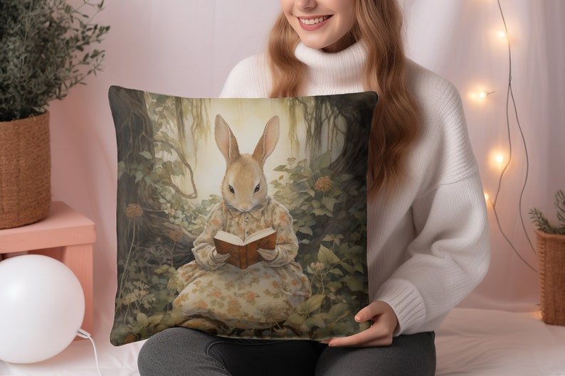 K&ouml;nnte beinhalten: Ein Dekokissen mit einer skurrilen Illustration eines Kaninchens, das ein Kleid tr&auml;gt und in einer Waldlandschaft ein Buch liest. Das Kissen hat eine braune, gr&uuml;ne und gelbe Farbgebung.