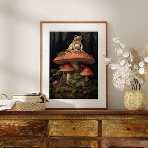 May include: A framed print of a frog sitting on a red and white mushroom in a forest setting. The mushroom is surrounded by other mushrooms and green foliage.