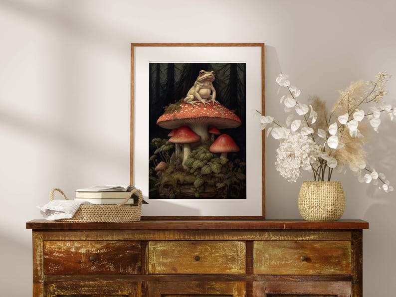 May include: A framed print of a frog sitting on a red and white mushroom in a forest setting. The mushroom is surrounded by other mushrooms and green foliage.