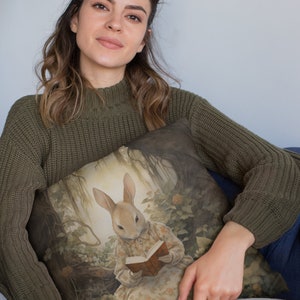 K&ouml;nnte beinhalten: Eine Frau in einem gr&uuml;nen Pullover h&auml;lt ein Kissen mit einem Gem&auml;lde eines Kaninchens, das in einer Waldlandschaft ein Buch liest.