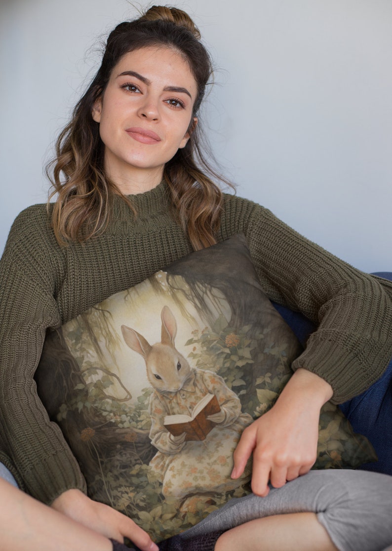 K&ouml;nnte beinhalten: Eine Frau in einem gr&uuml;nen Pullover h&auml;lt ein Kissen mit einem Gem&auml;lde eines Kaninchens, das in einer Waldlandschaft ein Buch liest.