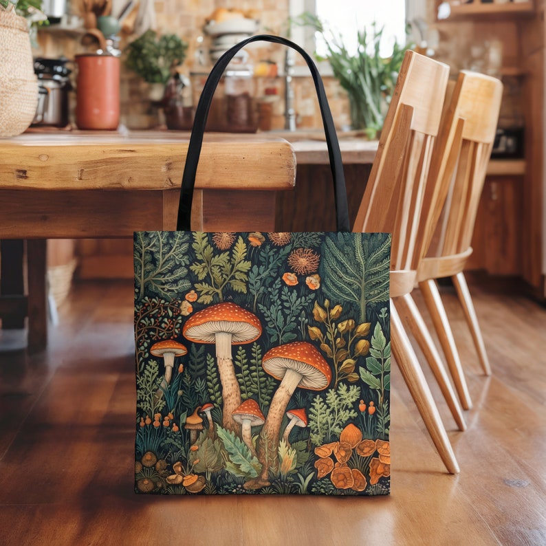 Puede incluir: Una bolsa de mano negra con una ilustraci&oacute;n colorida de setas y follaje. Las setas son rojas y naranjas con manchas blancas. El follaje es verde y marr&oacute;n.