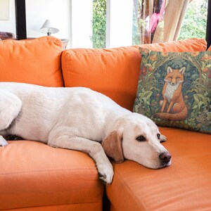 May include: A white dog with brown eyes is lying on an orange couch. A throw pillow with a fox illustration is on the couch behind the dog.