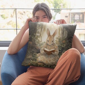 K&ouml;nnte beinhalten: Ein Dekokissen mit einer skurrilen Illustration eines Kaninchens, das ein Kleid tr&auml;gt und in einem Waldsetting ein Buch liest. Das Kissen ist in einem weichen, hellbraunen Farbton mit einem dezenten Blumenmuster gehalten.