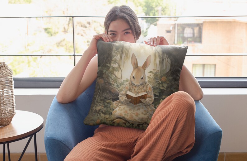 K&ouml;nnte beinhalten: Ein Dekokissen mit einer skurrilen Illustration eines Kaninchens, das ein Kleid tr&auml;gt und in einem Waldsetting ein Buch liest. Das Kissen ist in einem weichen, hellbraunen Farbton mit einem dezenten Blumenmuster gehalten.