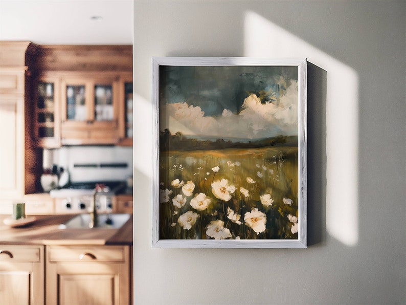 May include: A framed painting of a field of white wildflowers with a cloudy sky in the background. The painting is in a white frame and is hanging on a white wall.