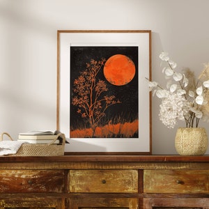 Peut inclure: Une impression encadr&eacute;e d'un arbre orange stylis&eacute; avec une grande lune orange en arri&egrave;re-plan. L'impression est plac&eacute;e contre un mur blanc avec une commode en bois et un vase de fleurs blanches au premier plan.