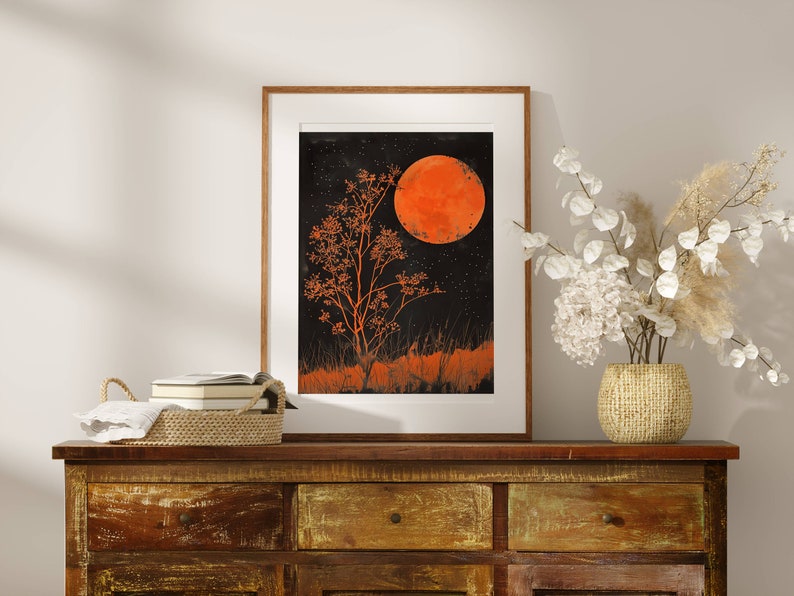 Peut inclure: Une impression encadr&eacute;e d'un arbre orange stylis&eacute; avec une grande lune orange en arri&egrave;re-plan. L'impression est plac&eacute;e contre un mur blanc avec une commode en bois et un vase de fleurs blanches au premier plan.