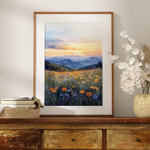 Puede incluir: Una pintura de acuarela enmarcada representa un paisaje de montaña al atardecer. La obra presenta un campo de flores silvestres en tonos naranja, amarillo y azul, con montañas azules onduladas al fondo. La pintura se exhibe sobre un mueble de madera.