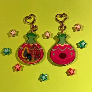 Puede incluir: Dos llaveros con un diseño rojo y verde, herrajes dorados y un cierre en forma de corazón. Un llavero tiene una escena interior detallada, el otro tiene un centro rojo liso. Alrededor hay adornos en forma de estrella.