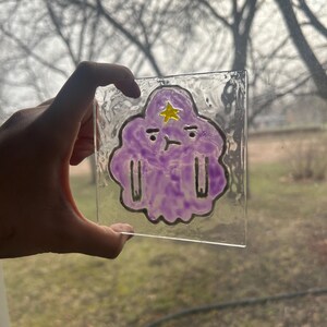 May include: A clear, textured glass square featuring a purple cloud-like character with a yellow star and black outlines. The character has a grumpy expression. The glass is held up against a blurred outdoor background.