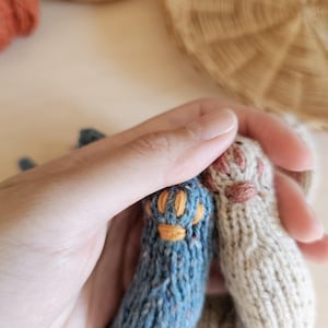 May include: Close-up of two knitted cat paws, one blue with orange details and the other beige with pink details. The paws are held in a hand, with yarn and a woven basket in the background. The knitted paws are likely part of a larger craft project.
