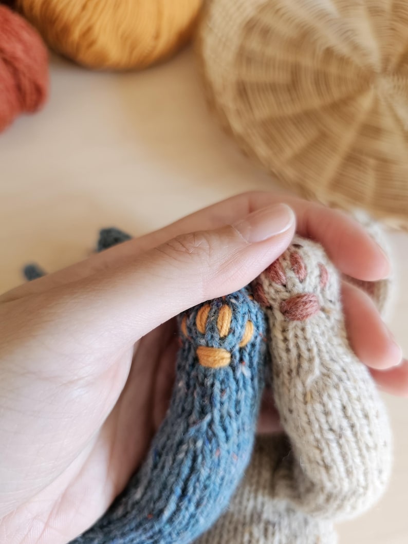 May include: Close-up of two knitted cat paws, one blue with orange details and the other beige with pink details. The paws are held in a hand, with yarn and a woven basket in the background. The knitted paws are likely part of a larger craft project.