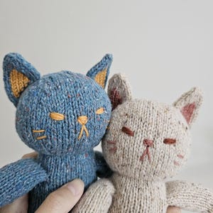 May include: Two knitted cat dolls. One is blue with orange ear lining and the other is beige with pink ear lining. Both have embroidered facial features, including closed eyes and a small nose.