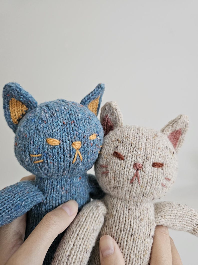 May include: Two knitted cat dolls. One is blue with orange ear lining and the other is beige with pink ear lining. Both have embroidered facial features, including closed eyes and a small nose.