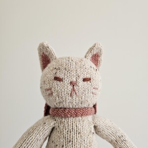 May include: A handmade knitted cat doll with a cream-colored body and a pink scarf. The cat has closed eyes, pink inner ears, and embroidered facial details. The doll's arms are outstretched, and it has a soft, textured appearance.