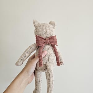 May include: A handmade, beige knitted cat doll with a textured appearance. The cat has a dark rose-colored bow tie. The doll has long legs and arms, and small, pointed ears. The cat is held by a person.