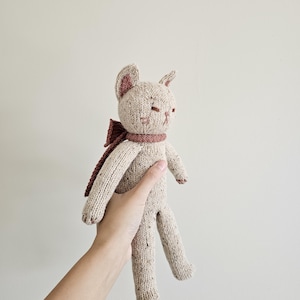 May include: A handmade knitted cat doll in beige with a brown scarf and bow. The cat has closed eyes and small ears. The doll is held in a hand against a plain background. The cat's paws and inner ears are a darker brown.