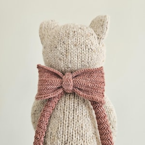 May include: A knitted teddy bear toy with a light beige body and a dusty rose bow. The bow is tied around the bear's neck and has a textured, woven appearance. The bear has small, pointed ears.