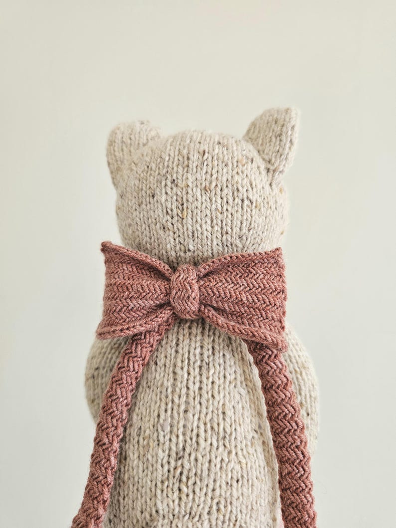 May include: A knitted teddy bear toy with a light beige body and a dusty rose bow. The bow is tied around the bear's neck and has a textured, woven appearance. The bear has small, pointed ears.