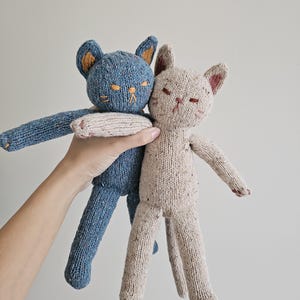 May include: Two knitted cat dolls. One is blue with orange ears and embroidered details, the other is beige with pink accents. Both have closed eyes and outstretched arms, suggesting a hug. The dolls are handmade and have a soft, textured appearance.