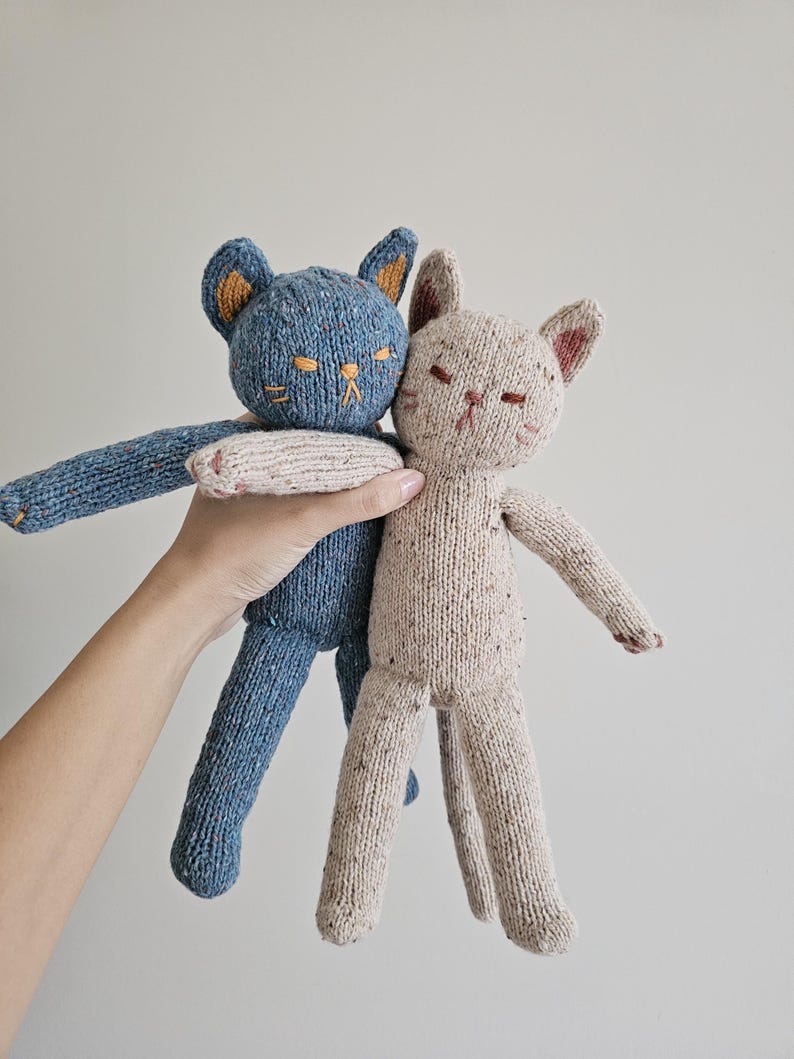 May include: Two knitted cat dolls. One is blue with orange ears and embroidered details, the other is beige with pink accents. Both have closed eyes and outstretched arms, suggesting a hug. The dolls are handmade and have a soft, textured appearance.