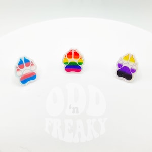 May include: Three paw print pins with different pride flag designs. The pins are white with a clear overlay and feature the transgender, rainbow, and non-binary pride flags.