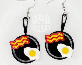 May include: A pair of novelty earrings featuring a black frying pan design with a fried egg and bacon. The earrings have silver-colored hooks and are set against a white background. The text "ODD FREAKY" is visible in the background.