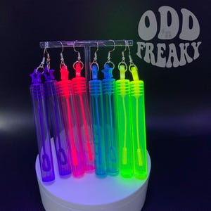May include: A set of seven colorful bubble wands, each with a silver hook, displayed on a white stand. The wands are neon purple, blue, pink, green, light blue, yellow, and green. The text "ODD FREAKY" is visible in the background.