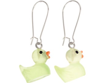 May include: A pair of drop earrings featuring translucent pale green rubber duck charms with small black eyes and bright orange beaks. The charms are suspended from silver-toned metal kidney-shaped ear wires, creating a playful and lightweight accessory.