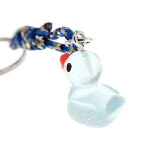 May include: A translucent light blue glass duck charm with a small red beak and black eyes, attached to a silver-toned metal jump ring and a blue, black, and white patterned braided cord that connects to a circular metal keychain ring.