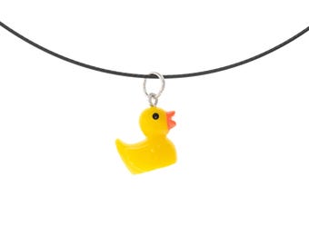 May include: A yellow rubber duck charm suspended from a thin black cord. The duck features a bright yellow body, a small orange beak, and a single black eye. The charm is attached to the cord by a small silver-toned metal jump ring.