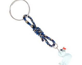 Peut inclure: Un porte-clés composé d'un anneau en métal argenté relié à une cordelette nouée au motif tressé bleu, noir et beige. La cordelette est fixée à une petite breloque en forme de canard bleu clair translucide avec un bec orange vif.