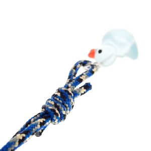 May include: A braided rope lanyard featuring a blue, black, and grey patterned cord tied in a knot. Attached to the end of the cord is a small, light blue duck-shaped charm with a bright orange beak and a single black eye, connected by a metal ring.