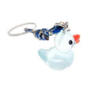 May include: A keyring featuring a translucent light blue glass duck charm with a bright orange beak and black eyes. The charm is attached to a braided cord in shades of blue, black, and tan, which connects to a circular silver metal key ring.