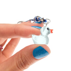 May include: A small, translucent white rubber duck charm with a bright orange beak and black eyes. The duck is attached to a silver-toned metal jump ring, which is connected to a braided cord featuring blue, black, and tan woven patterns.