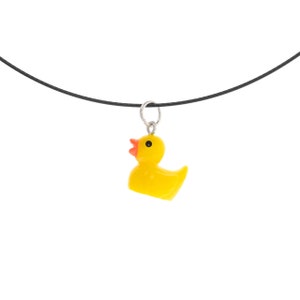 Könnte beinhalten: Ein gelber Gummi-Enten-Anhänger mit orangefarbenem Schnabel und schwarzem Auge, der an einem silberfarbenen Metallring an einer dünnen schwarzen Kordelkette hängt. Die Ente ist im Profil nach links ausgerichtet und hat eine glatte, glänzende Oberfläche.