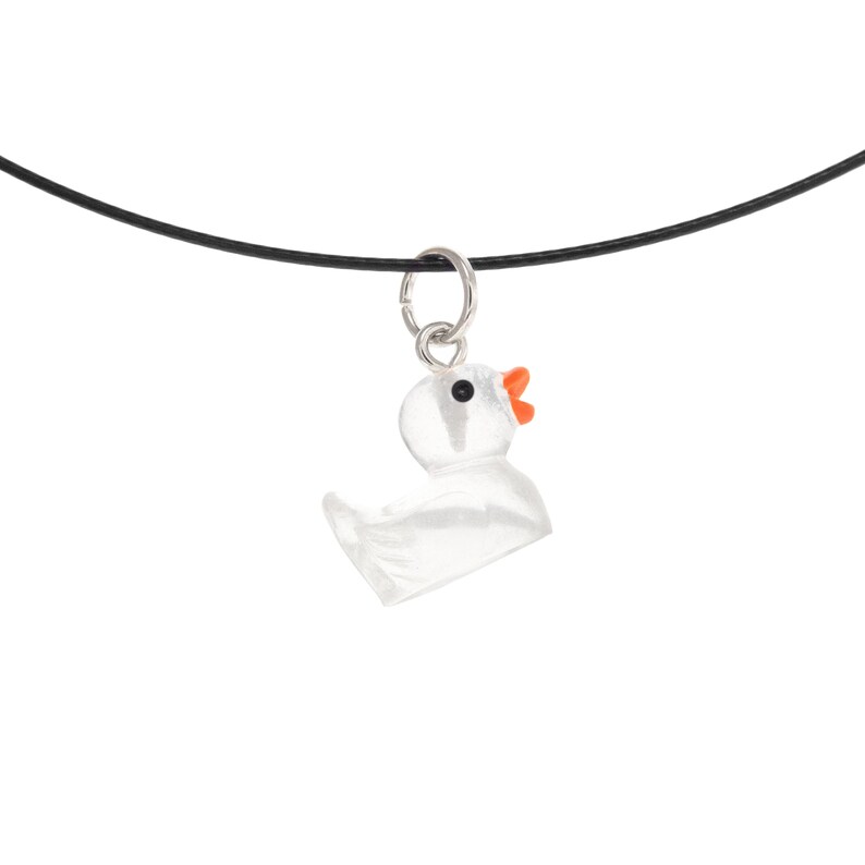 May include: A clear, translucent duck-shaped pendant hanging from a thin black cord. The duck features a small orange beak and a single black eye, attached to the cord by a silver-toned metal jump ring, creating a simple necklace design.