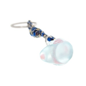 May include: A keyring featuring a translucent, pale blue glass charm with soft pink inclusions, attached to a silver-toned metal ring by a braided cord in shades of deep blue, black, and metallic gold thread, creating a textured, multi-material accessory.