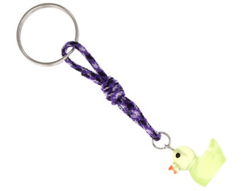 May include: A keyring featuring a small, pale yellow rubber duck charm with a bright orange beak and a black eye. The charm is attached to a braided cord in shades of purple, black, and white, which is connected to a circular silver metal ring.