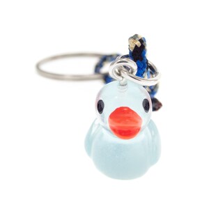 May include: A keyring featuring a small, translucent light blue duck charm with a bright orange beak and black eyes. The duck is attached to a silver-toned metal split ring by a short, braided cord in shades of blue, black, and tan.