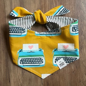 May include: A yellow bandana with a black and white striped trim. The bandana features a repeating pattern of vintage blue typewriters with a pink heart on a piece of paper. The bandana has a bow tie at the top and the text "Swoon Studio" in a white circle.