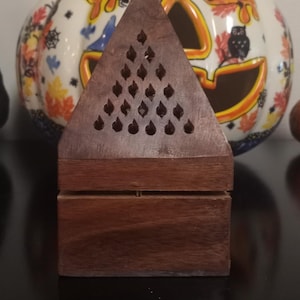 May include: A wooden triangular incense burner with a decorative pattern of small holes. The burner is made of dark brown wood and has a two-tiered design.