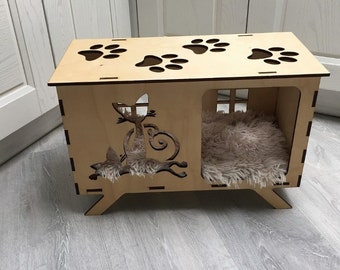 Op de afbeelding: Een houten kattenbed met een uitsnijding van twee katten en potafdrukken op de bovenkant. Het bed heeft een zacht, bruin kussen erin en staat op vier poten.