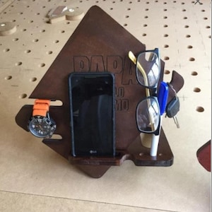 May include: A wooden stand with the word "DADA" engraved on it. The stand has slots for a phone, glasses, a watch, and keys.