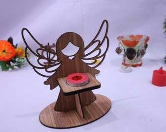 Peut inclure: Bougeoir en forme d'ange en bois avec une petite bougie rouge. L'ange a des ailes découpées et tient une plateforme carrée pour la bougie. La base est ovale. D'autres bougies et un verre décoratif sont en arrière-plan.